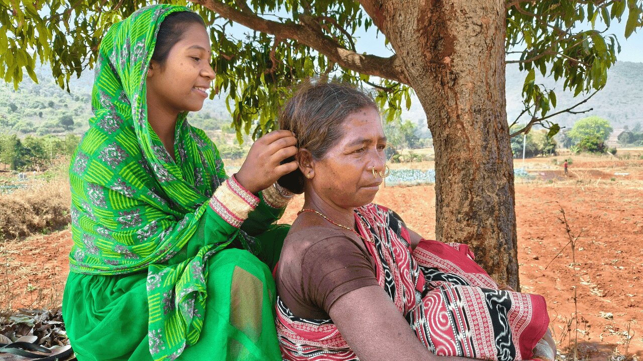 A young woman and an older woman sitting outside under a tree the younger woman is tending to the older womans hair