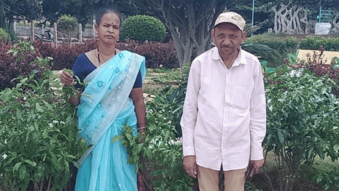An image of a woman and a man standing outside in front of a hedge looking at the camera