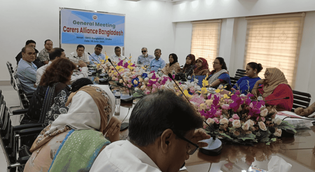 Big group of poeple sitting inside around a large conference table. Banner reading Carers Alliance Bangladesh hanging on the wall