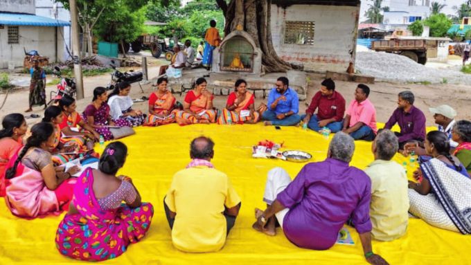 Image of large group of poeple sitting in a circle on the floor outside on a bright yellow tarpaulin