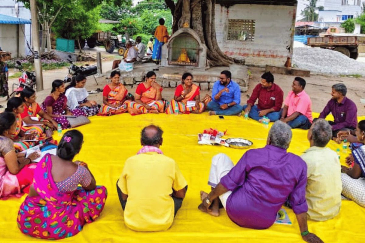 Image of large group of poeple sitting in a circle on the floor outside on a bright yellow tarpaulin