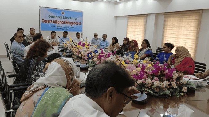 Big group of poeple sitting inside around a large conference table. Banner reading Carers Alliance Bangladesh hanging on the wall
