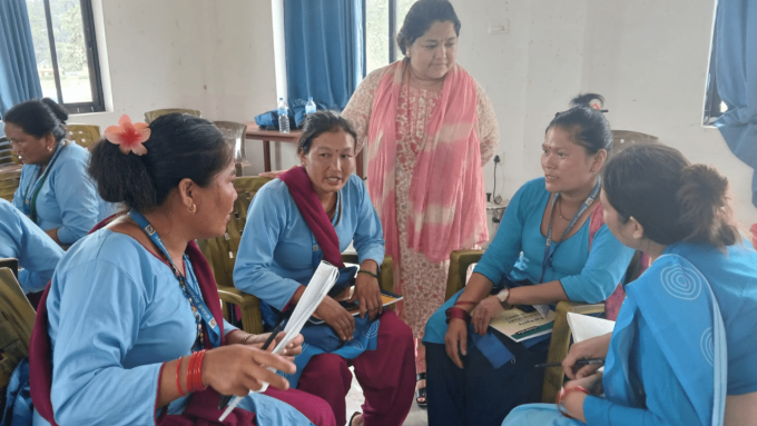 A small group of women sitting in a circle indoors talking with one woman standing beside them listening