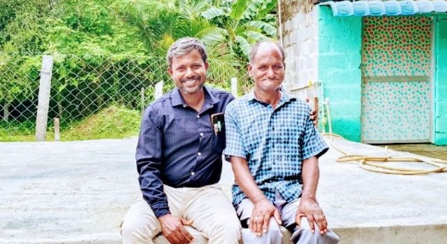 Two men sitting on a cement wall in front of a house, engaged in conversation and enjoying the outdoors.